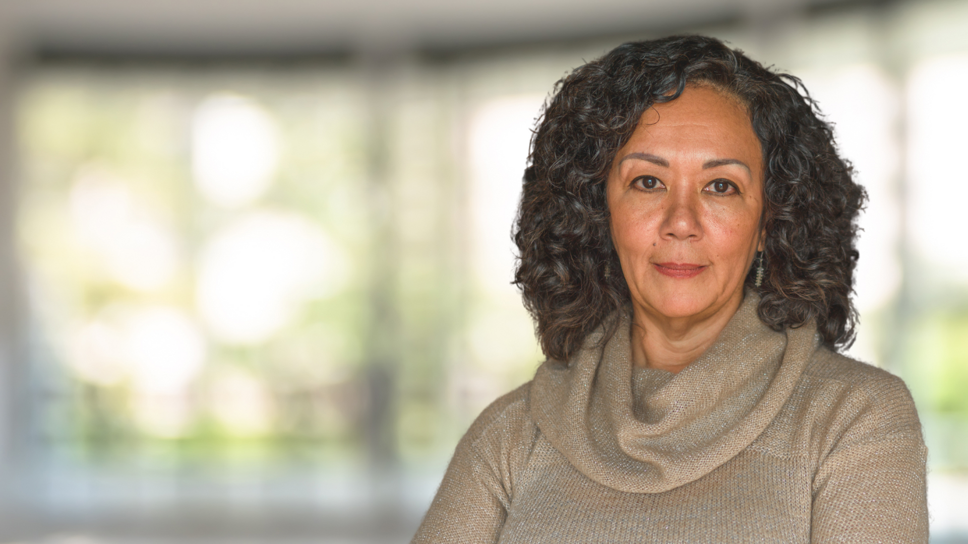 A professional woman with curly hair is wearing a sweater and standing in a hallway.