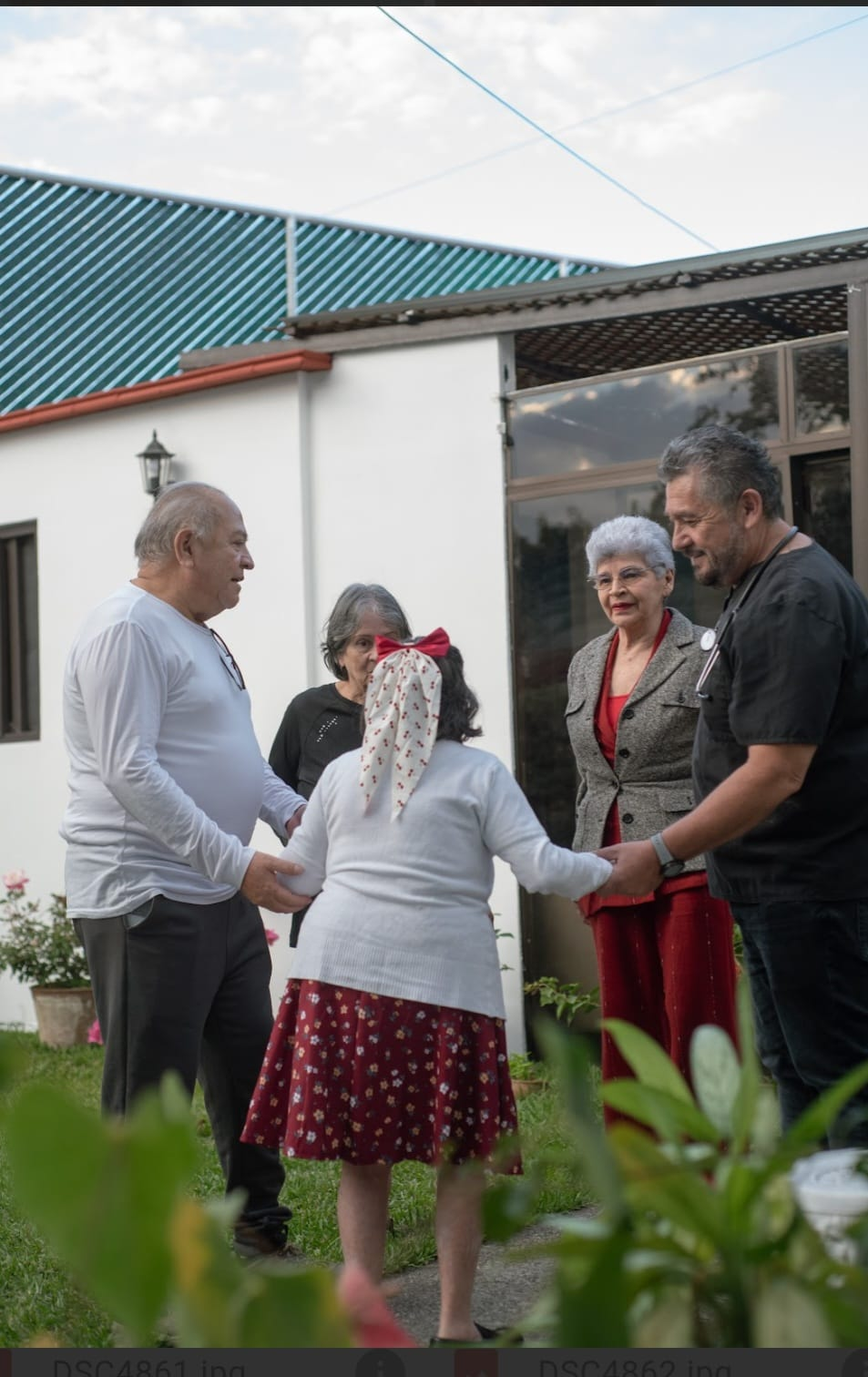 Cinco personas de pie en el patio de un jardín, tomadas de la mano y conversando cerca de la entrada de un edificio.
