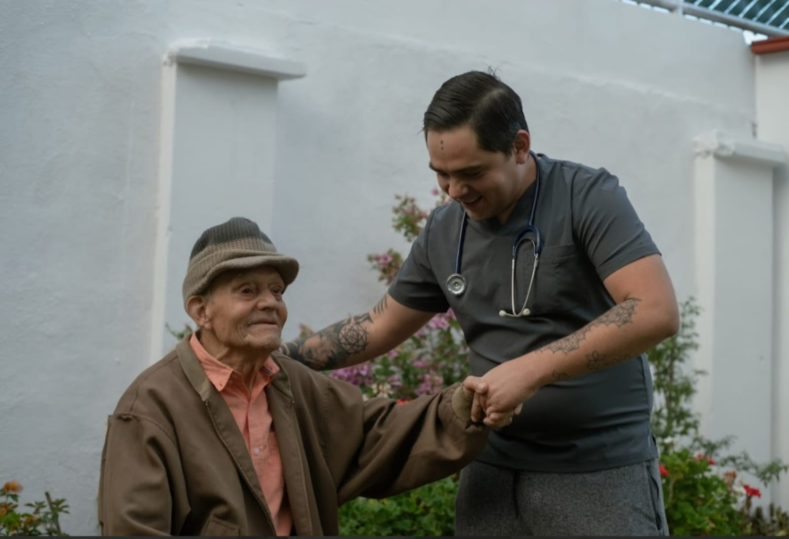 Un hombre ayuda a un hombre mayor con sombrero al aire libre junto a una pared blanca; ambos sonríen.