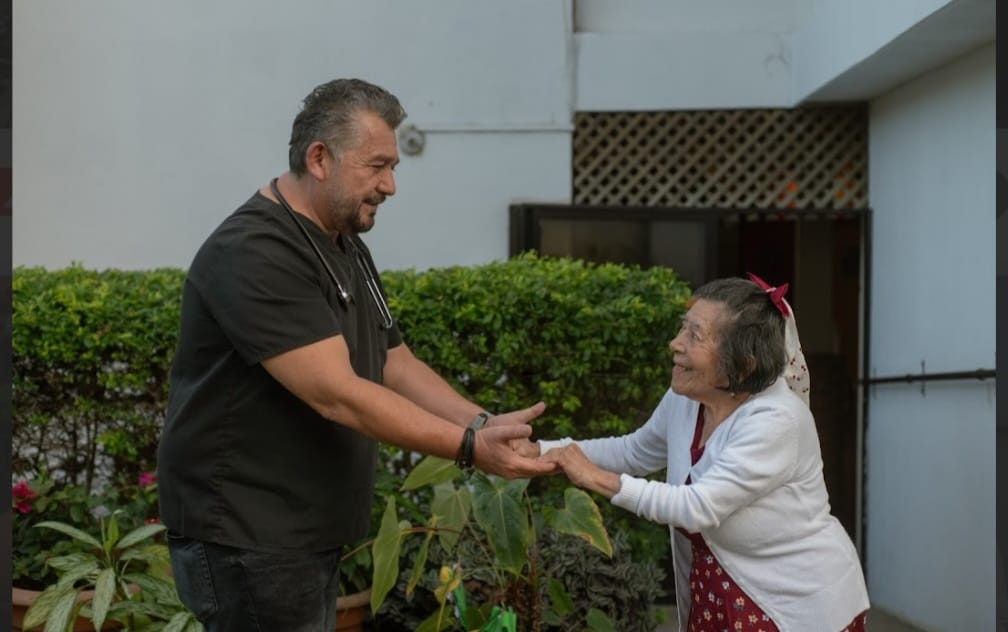 Dos personas tomadas de la mano y sonriendo al aire libre junto a un edificio y vegetación.