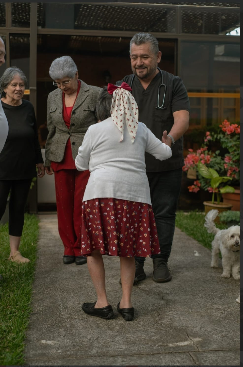 Varias personas charlan al aire libre junto a un edificio, con un perrito blanco cerca.