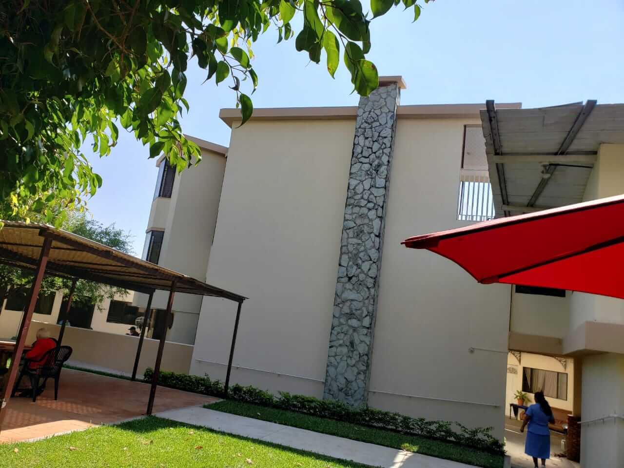 Patio con un edificio de color beige, una columna de piedra, sombrillas de patio y una persona caminando cerca de la entrada.
