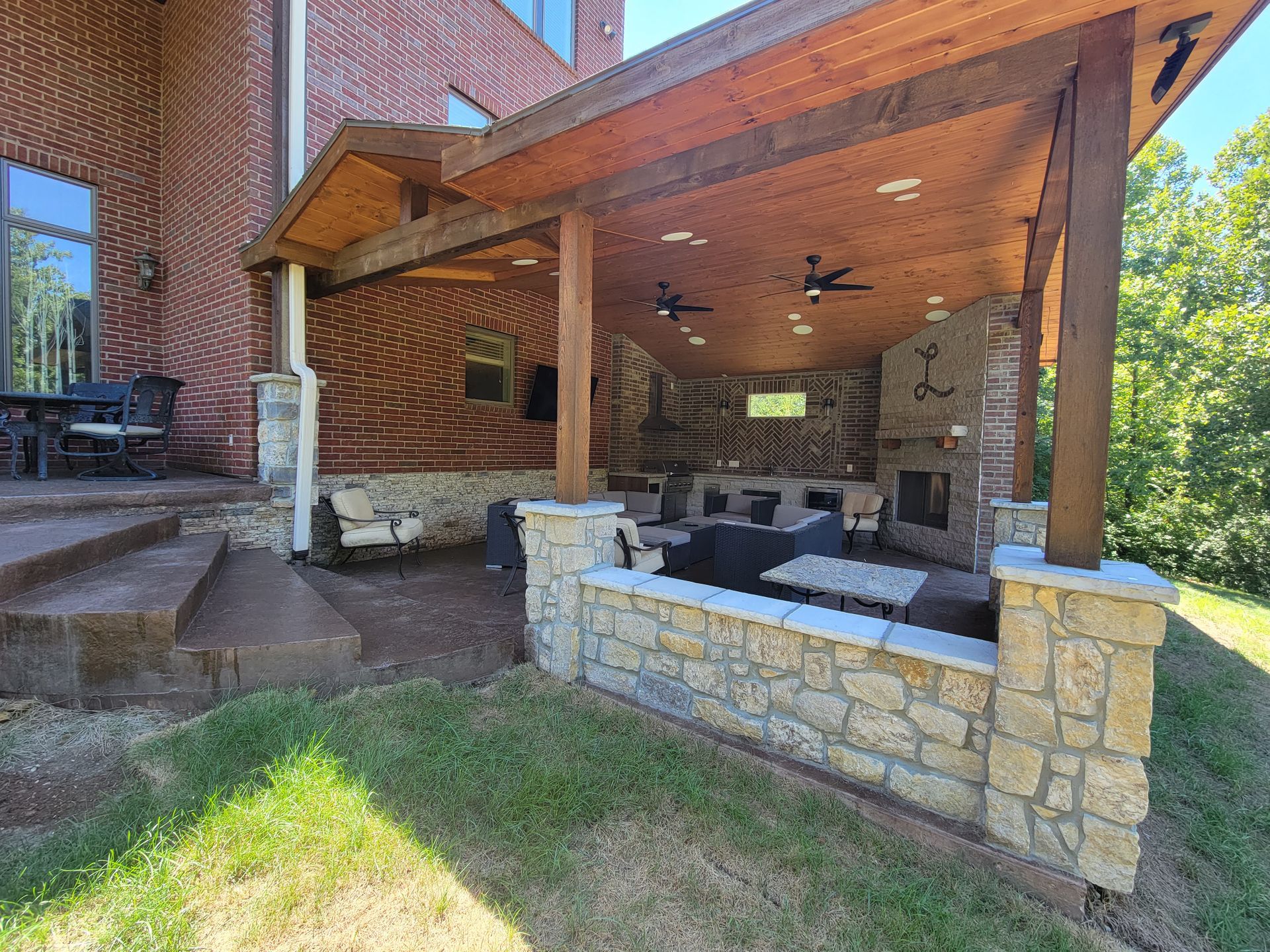 Covered stone patio with wood beams, outdoor seating, and brick house exterior beside a grassy yard