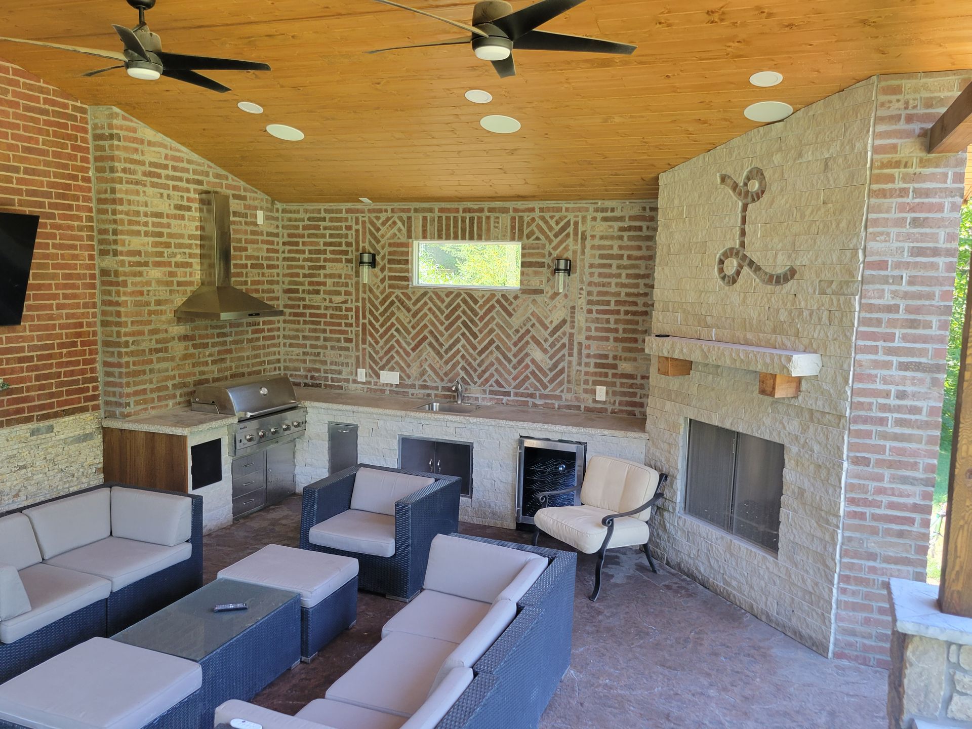 Covered patio with brick walls, cushioned seating, ceiling fans, and a fireplace around a TV.