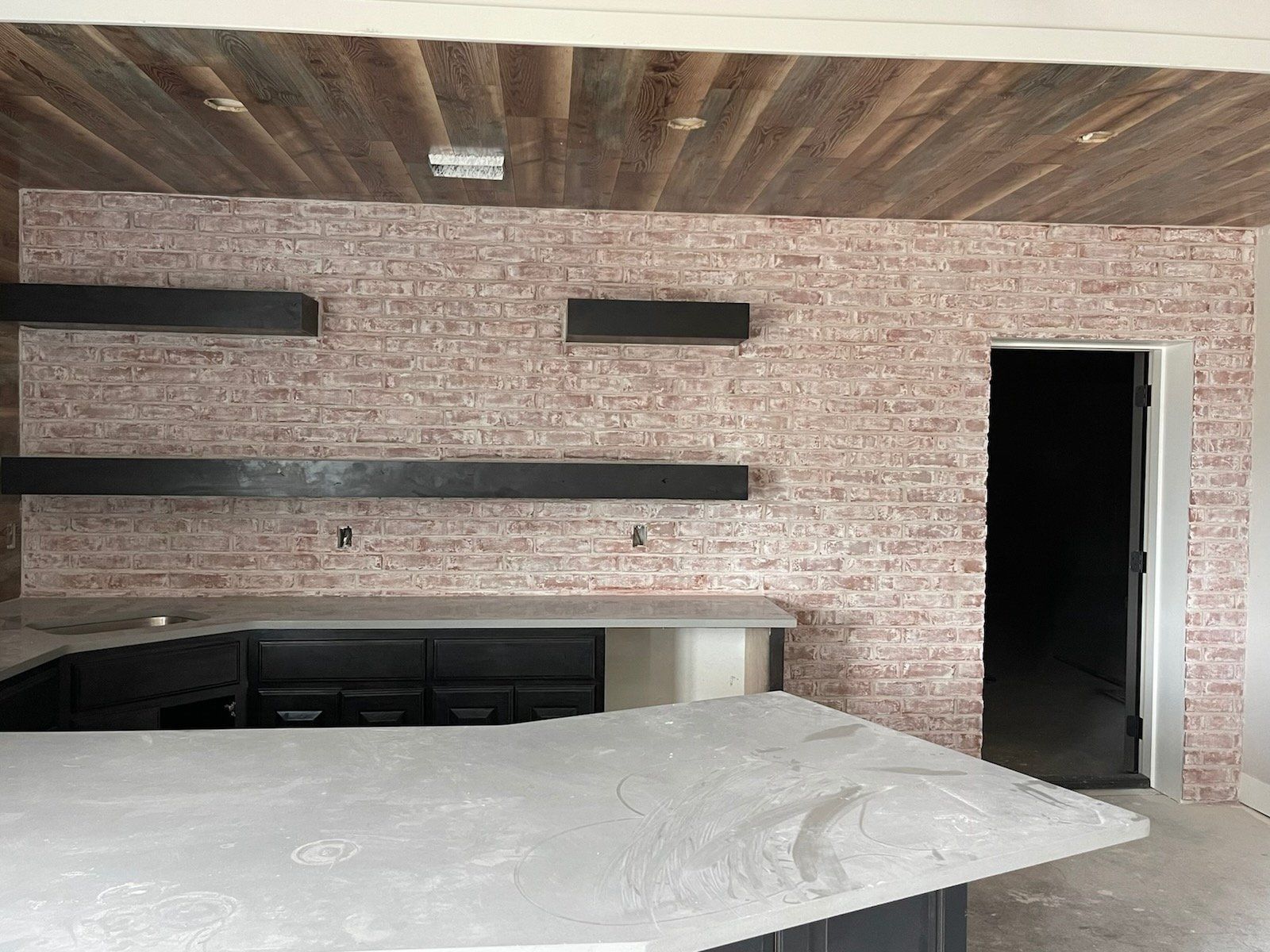Modern kitchen with white island, black cabinets, brick wall, wood ceiling, and open doorway