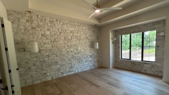 Empty living room with wood floors, stone accent wall, ceiling fan, and large window.