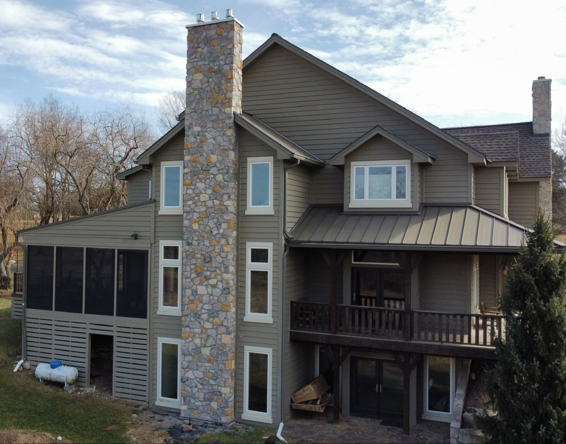 Stone-and-wood house exterior with a large chimney, screened porch, and deck under a cloudy sky