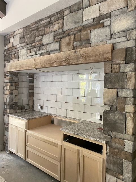 Stone fireplace surround with white tile backsplash and unfinished wooden cabinets in a kitchen area