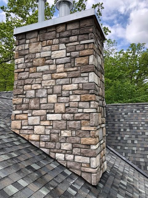 Stone chimney rising from a shingled roof with leafy trees in the background