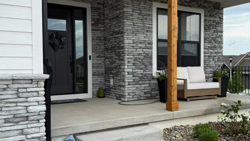 Front porch with gray stone siding, white columns, black door, and cushioned bench on a concrete patio.
