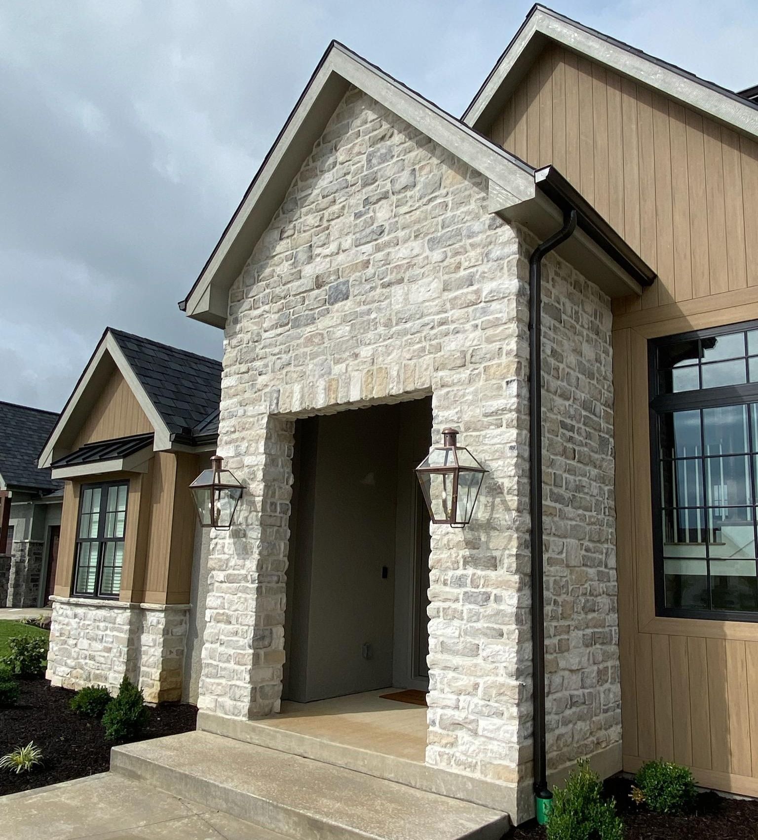 Stone house entryway with a gabled roof, tan siding, black trim, and front steps under a cloudy sky