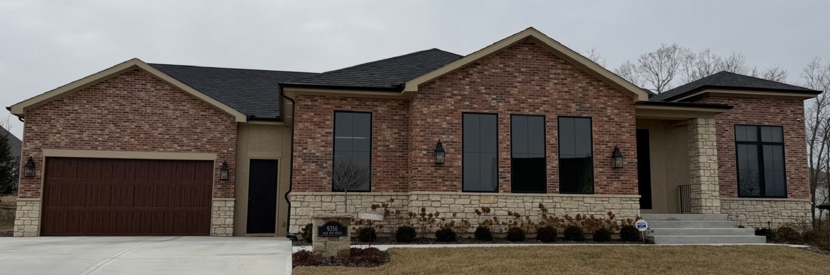 Single-story brick house with a two-car garage, stone accents, and dark roof under an overcast sky