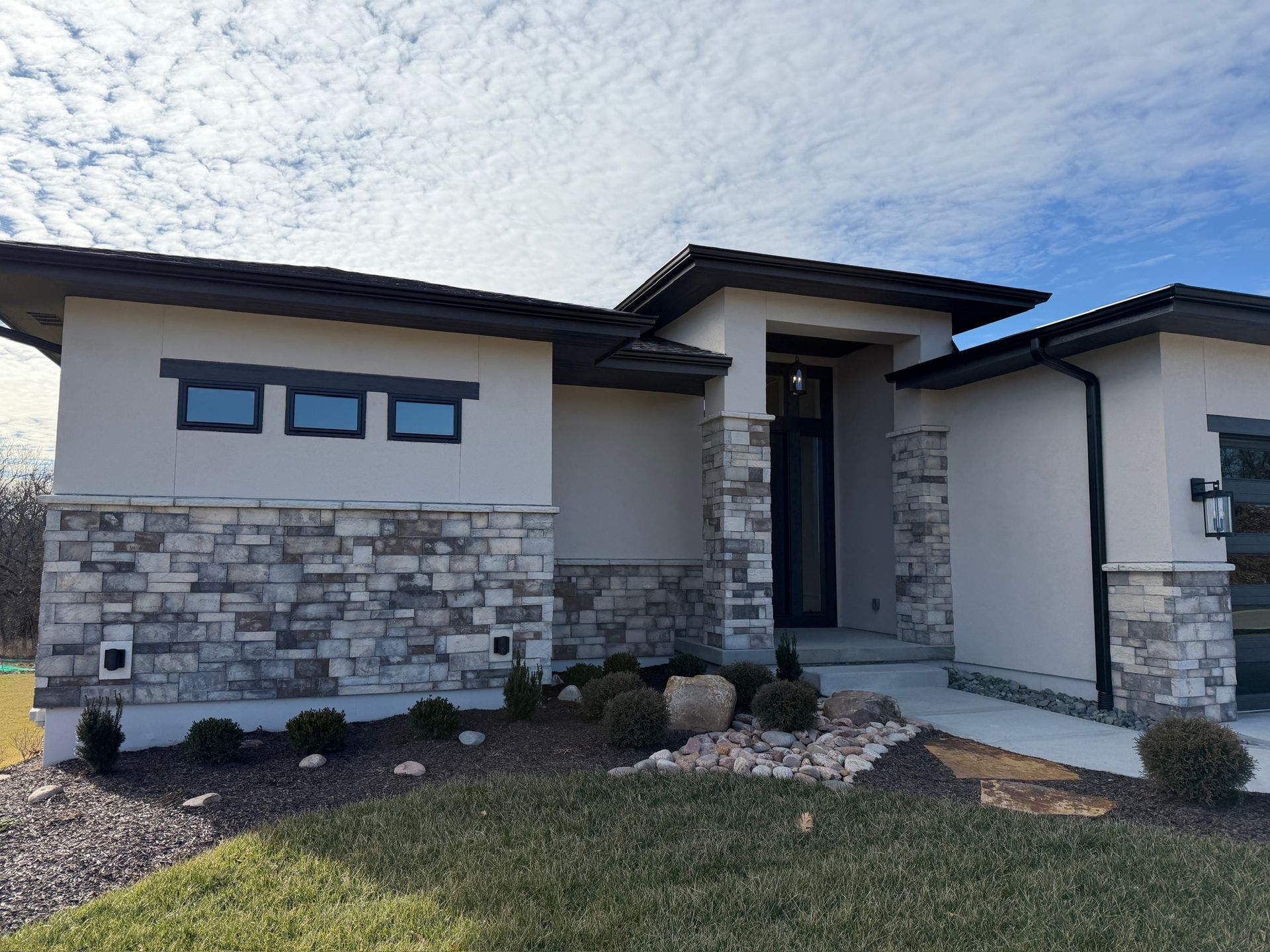 Modern single-story house exterior with stone accents, front entry, and landscaped yard under a blue sky