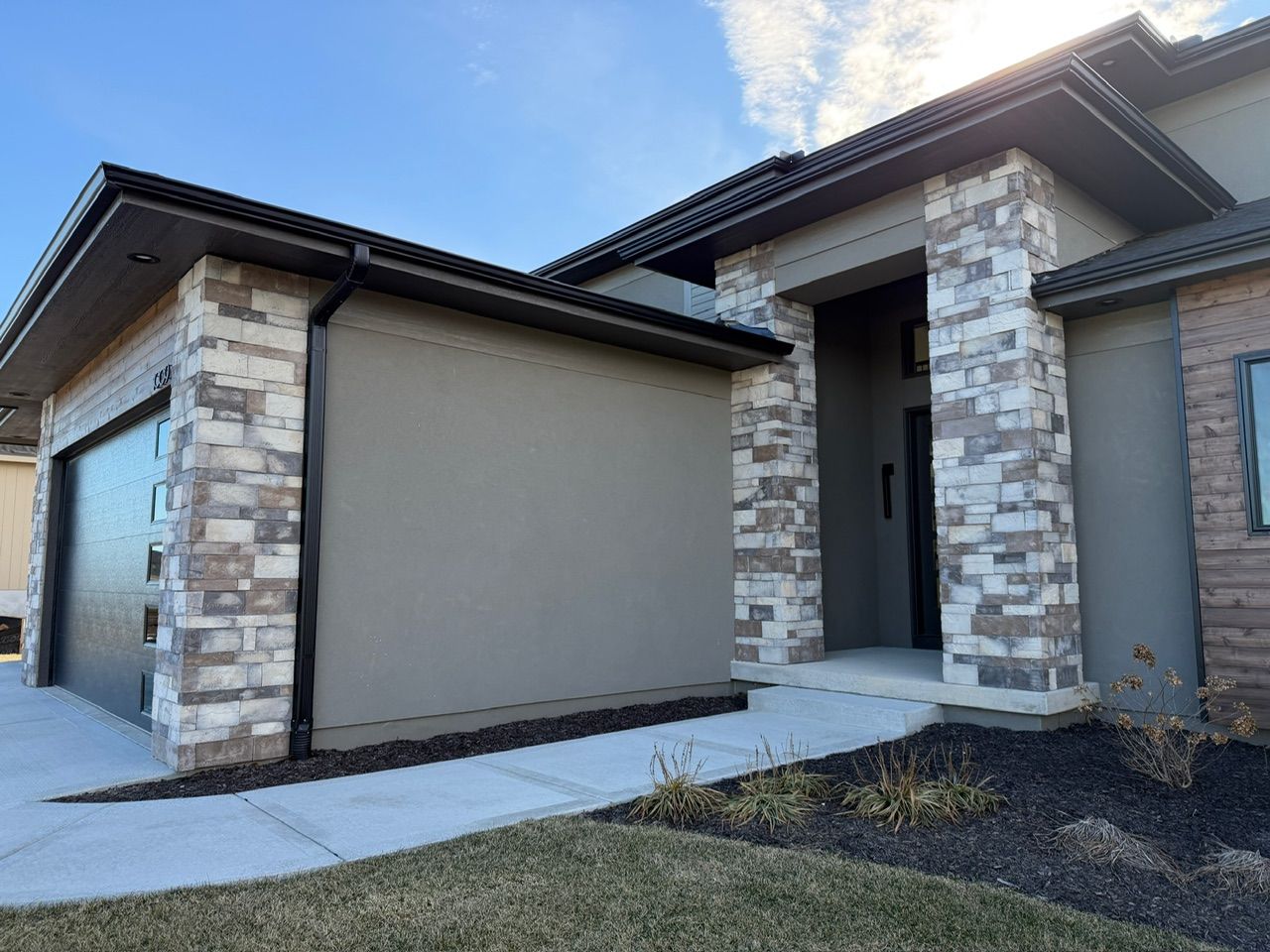 Modern house exterior with stone columns, beige walls, and a concrete walkway under a blue sky