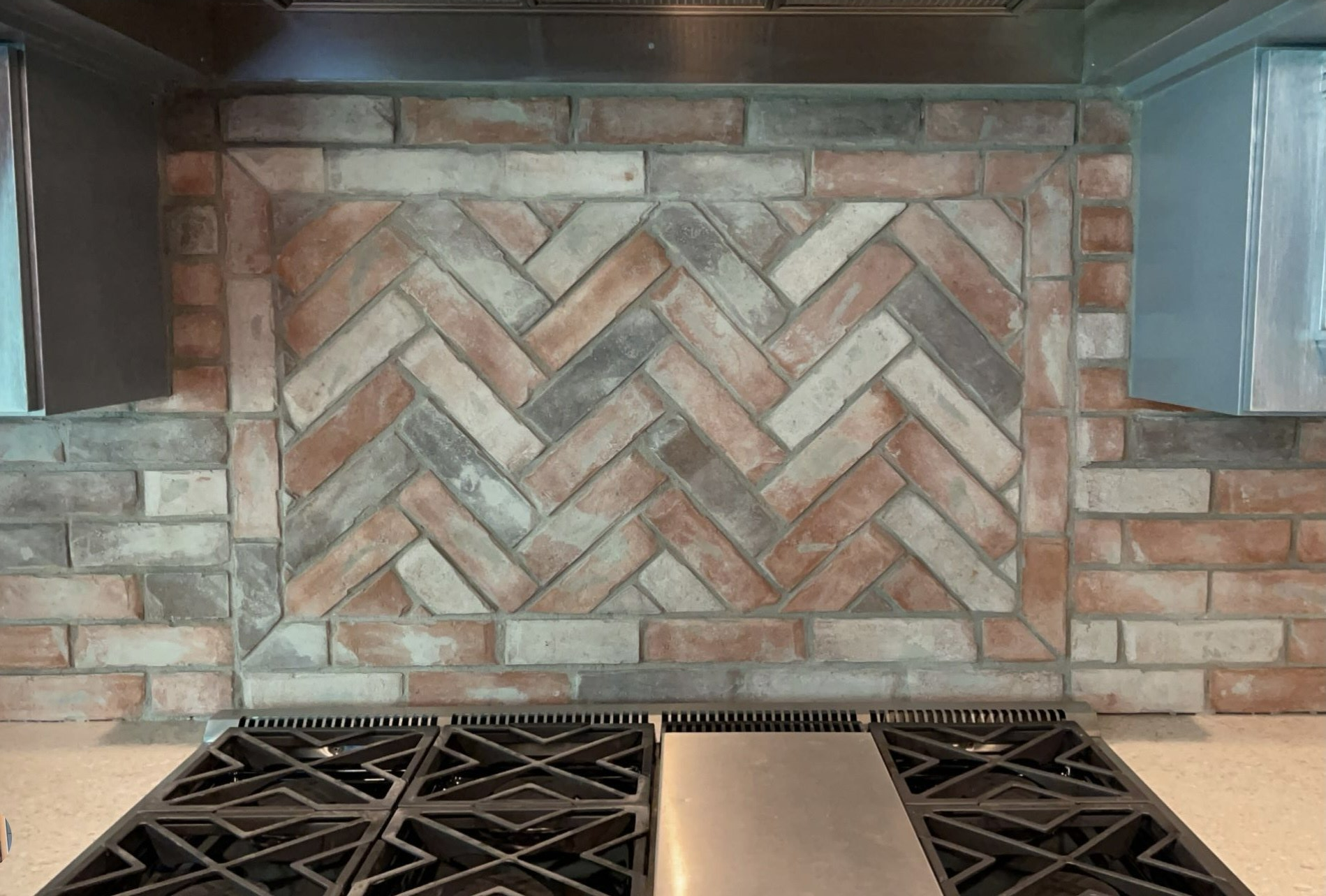 Brick backsplash behind a stovetop, with a herringbone tile panel and dark range burners below