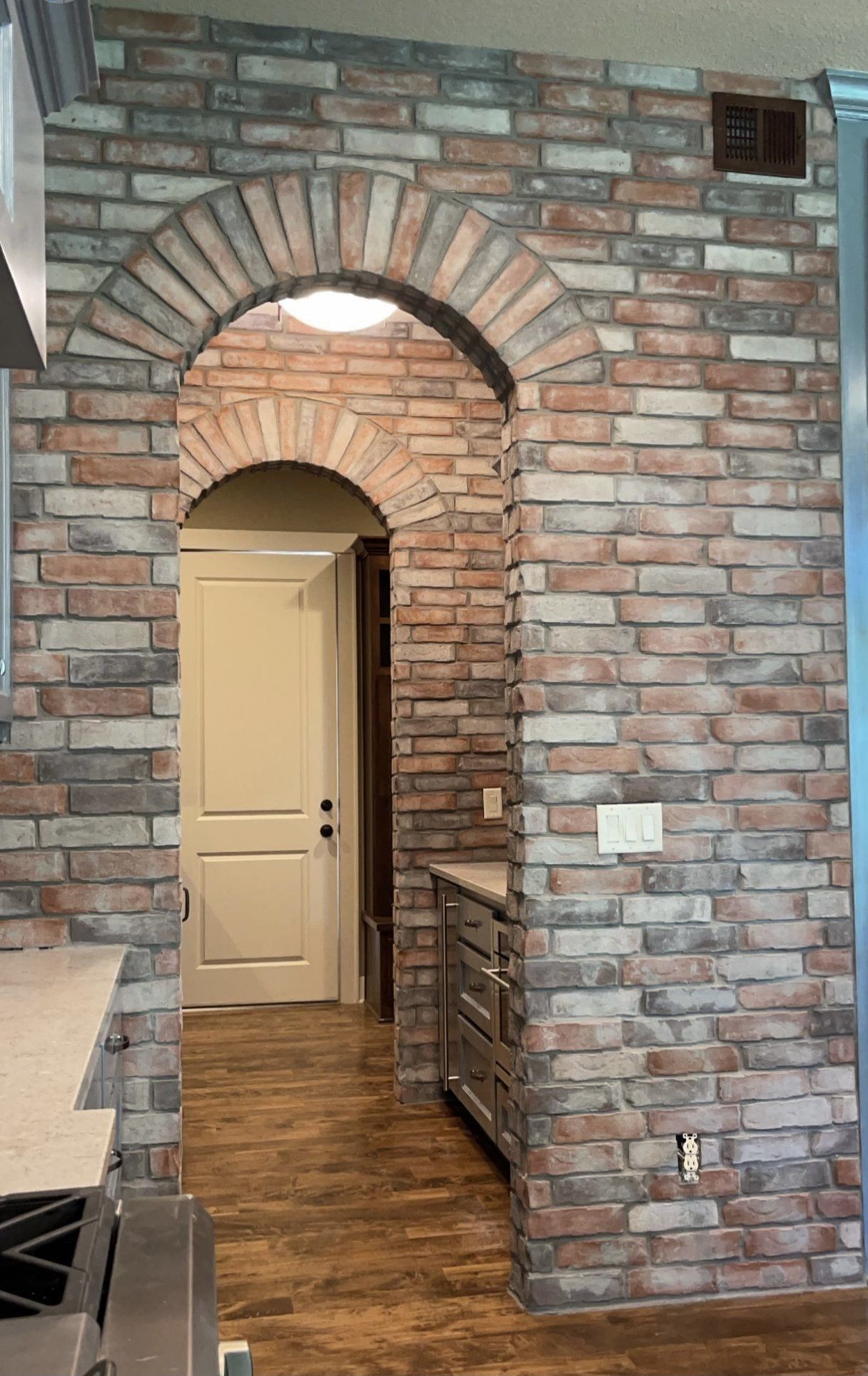 Brick interior hallway with arched doorway and open beige door, wood floor, and metal shelving on the right