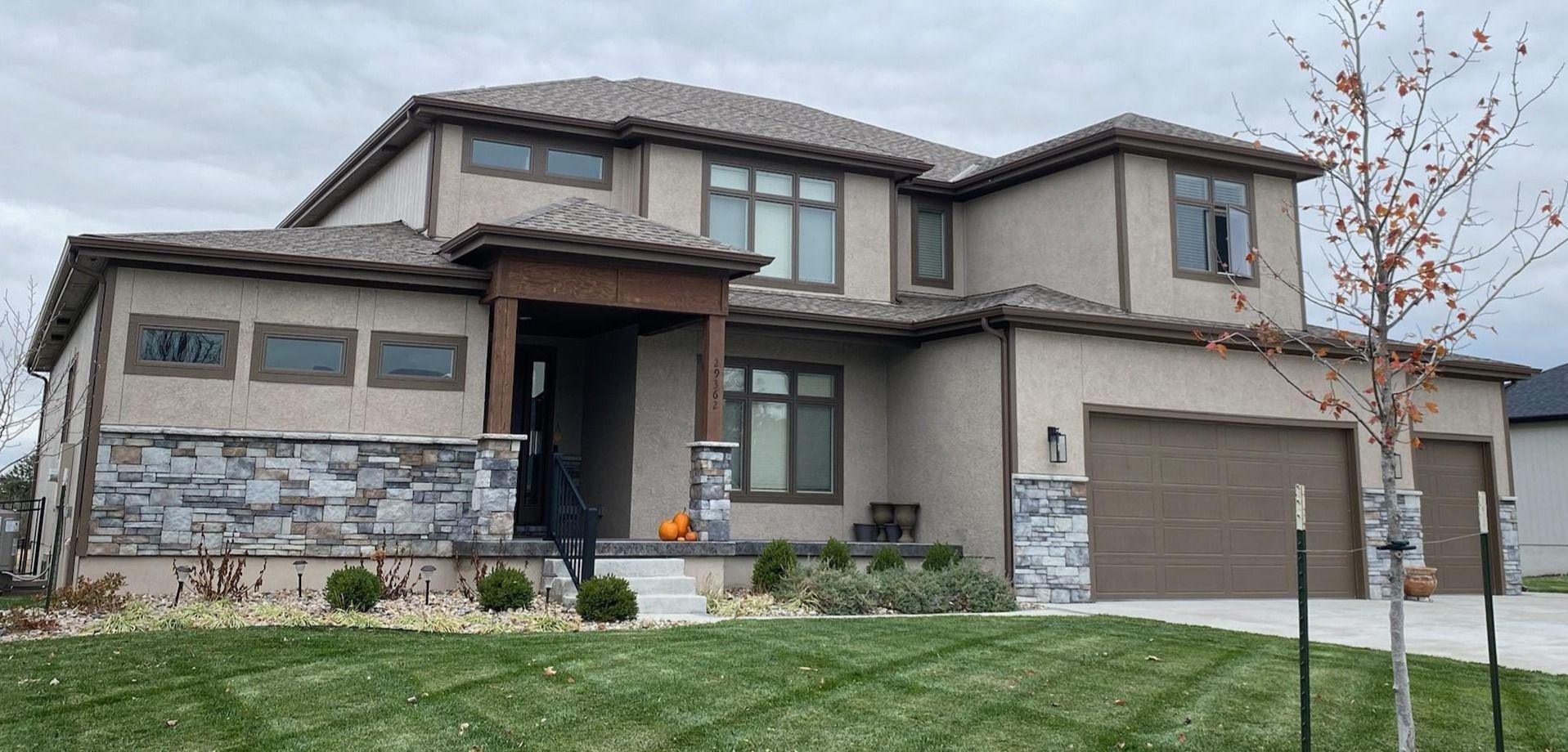 Two-story beige house with stone accents, front lawn, and attached garage under an overcast sky