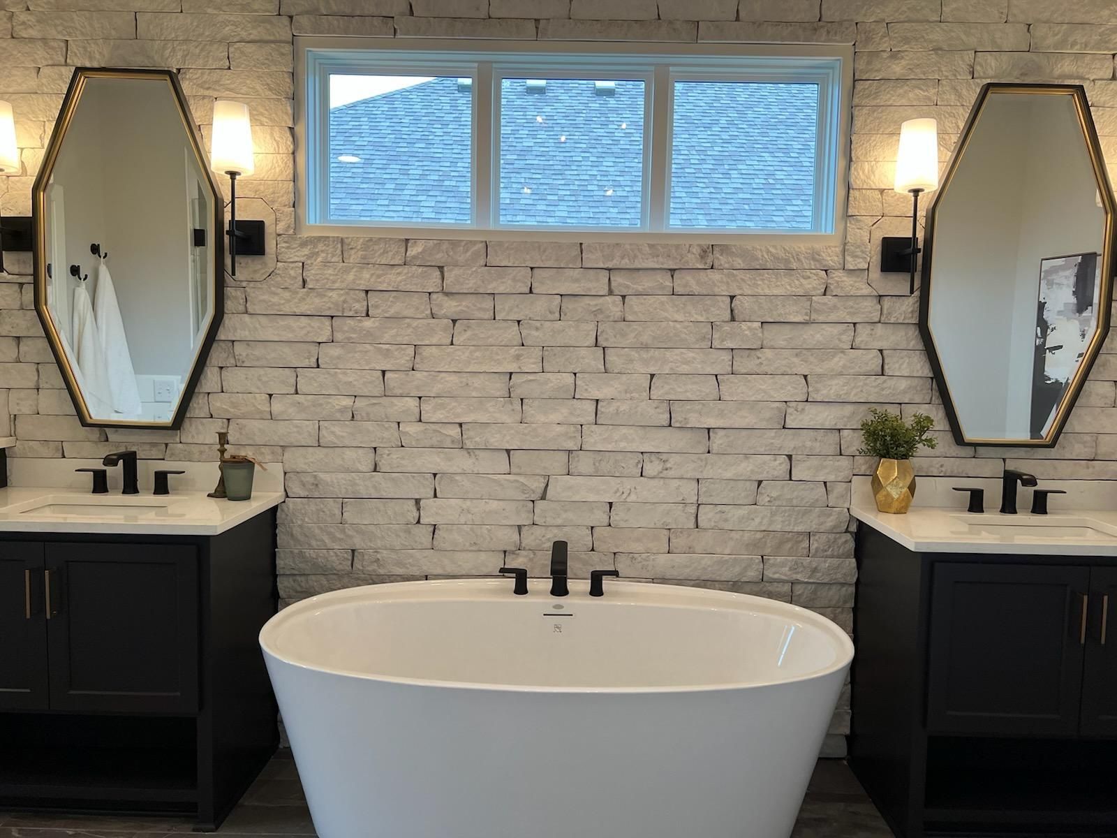 Modern bathroom with freestanding tub, black vanities, and a gray brick accent wall with windows and sconces