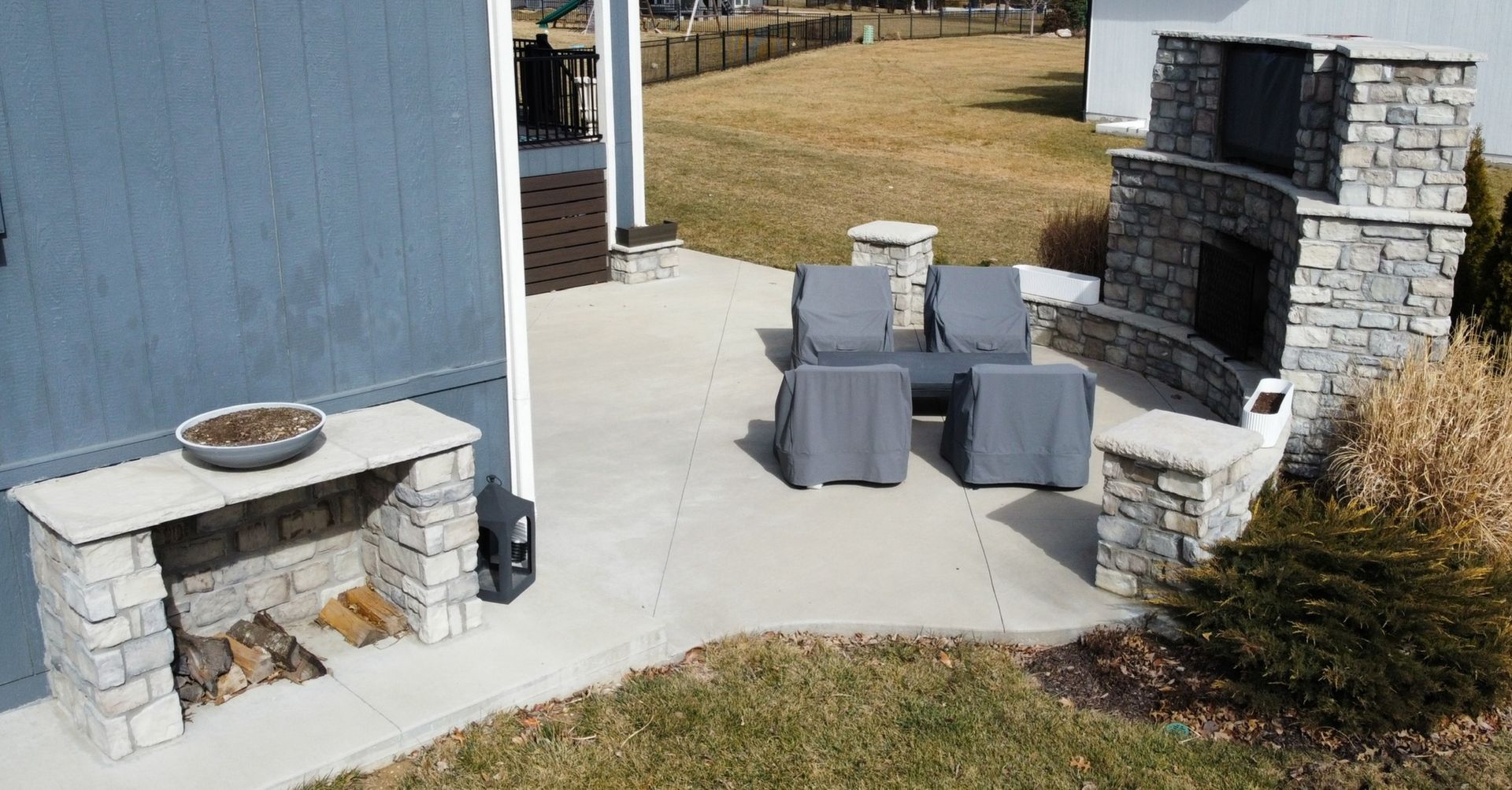 Patio with two gray chairs, a fire pit, and a stone outdoor fireplace beside a house