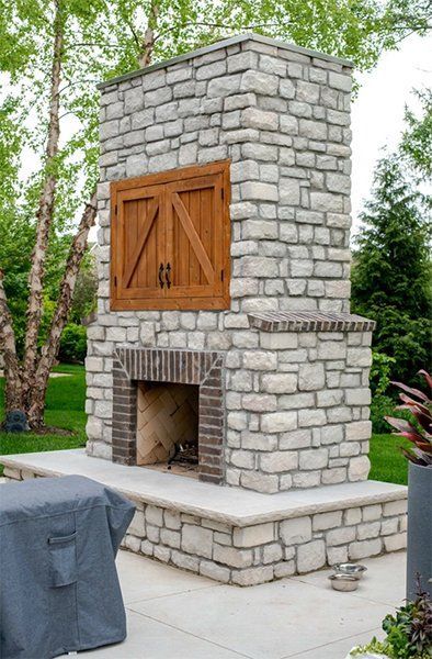 Stone outdoor fireplace with wooden cabinet door on a patio, surrounded by trees and greenery