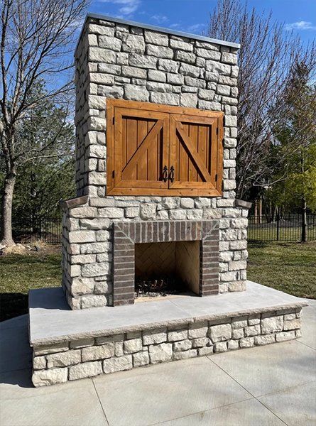 Stone outdoor fireplace with wooden doors on a concrete patio in a park-like setting