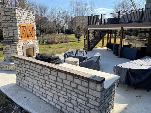 Stone patio with outdoor fireplace, covered lounge seating, and an elevated deck in a backyard