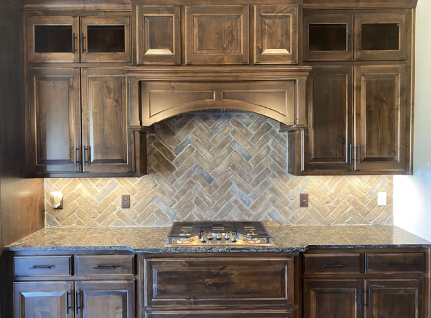 Dark wood kitchen cabinets with beige herringbone tile backsplash and stove beneath a hood vent.