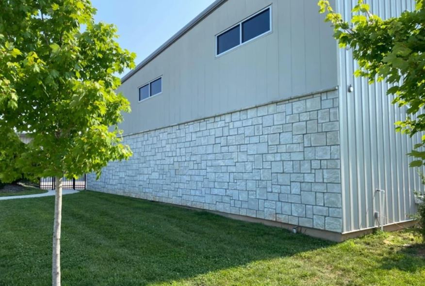 White industrial building with stone lower wall, blue windows, and green trees on a grassy slope