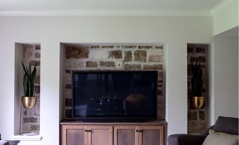 Living room wall with mounted TV above a wooden cabinet, framed by stone and two narrow plant alcoves