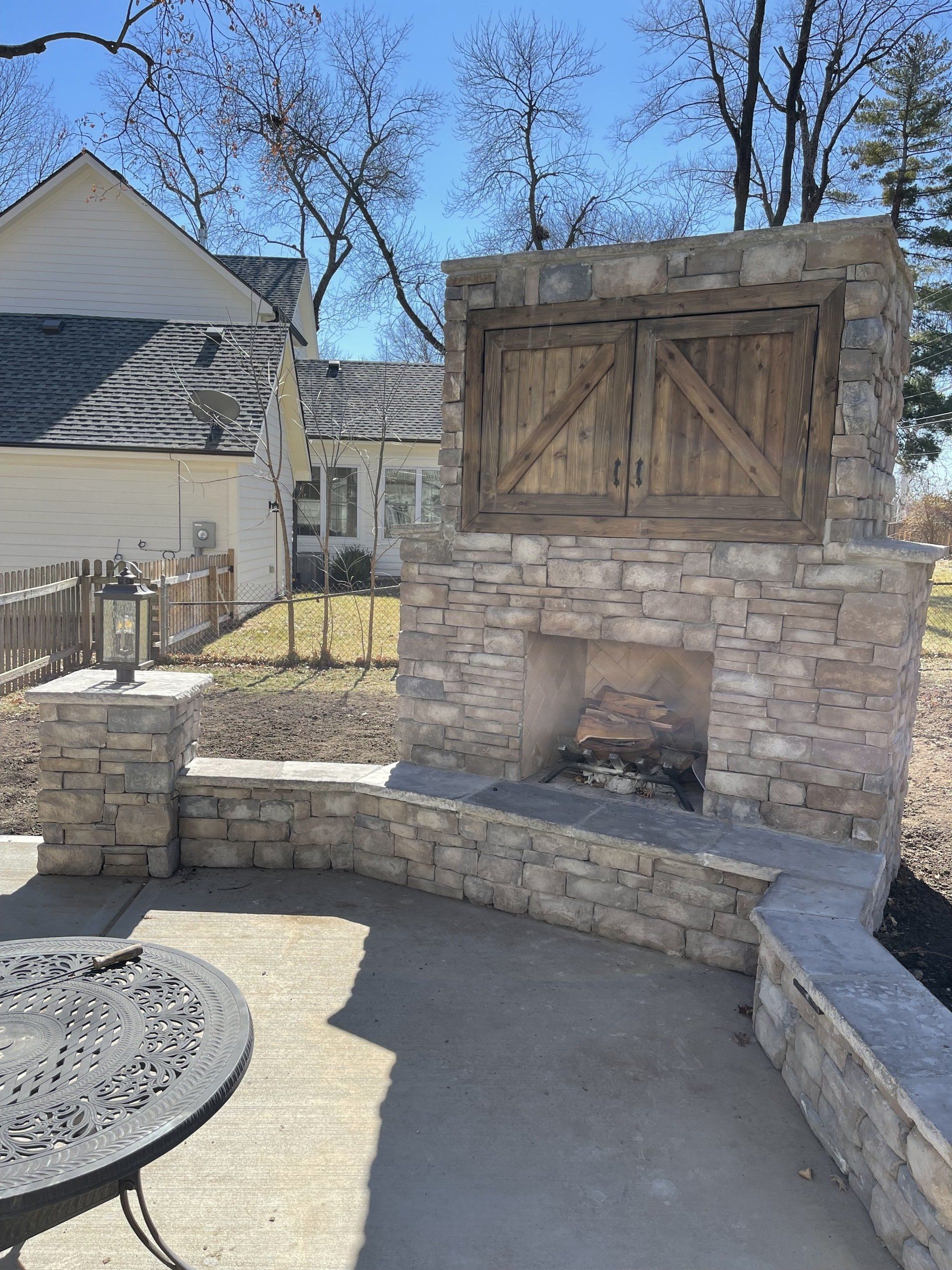 Stone outdoor fireplace with wooden doors, curved bench seating, and a round table in a sunny patio.
