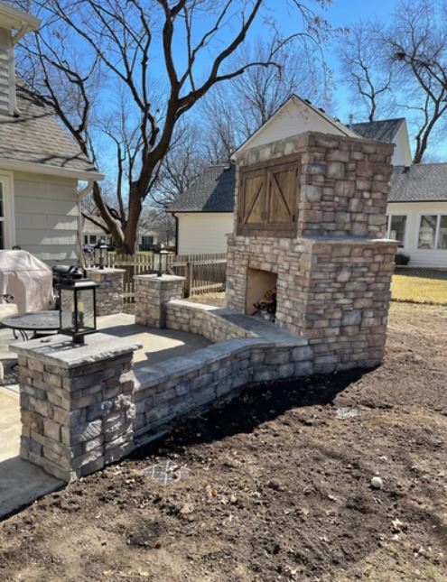 Stone outdoor fireplace and seating wall in a backyard with houses and bare trees in the background