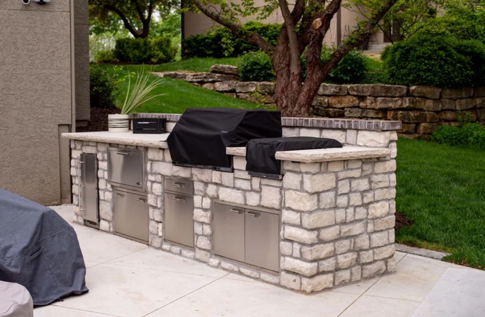 Stone outdoor kitchen with built-in grill and cabinets on a patio, with lawn and trees behind it