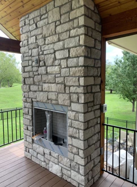 Stone outdoor fireplace on a covered porch with wooden ceiling and black railing overlooking a green yard