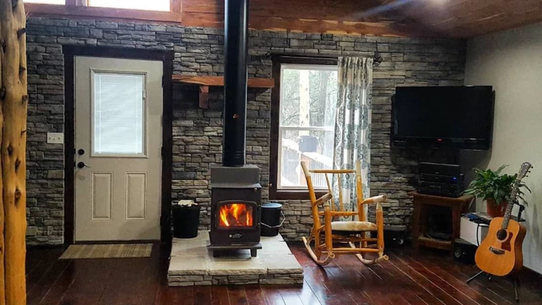 Cozy cabin interior with wood stove, rocking chair, door, windows, TV, and guitar by the wall