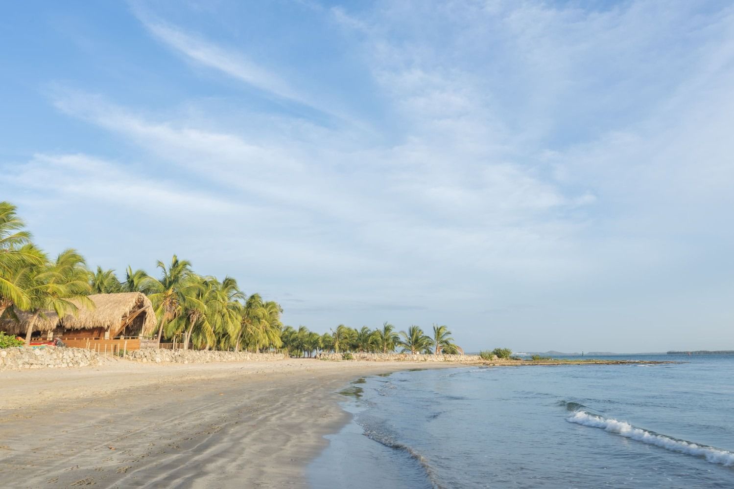 Beach with calm water, tan sand, palm trees, and thatched-roof buildings under a blue sky.
