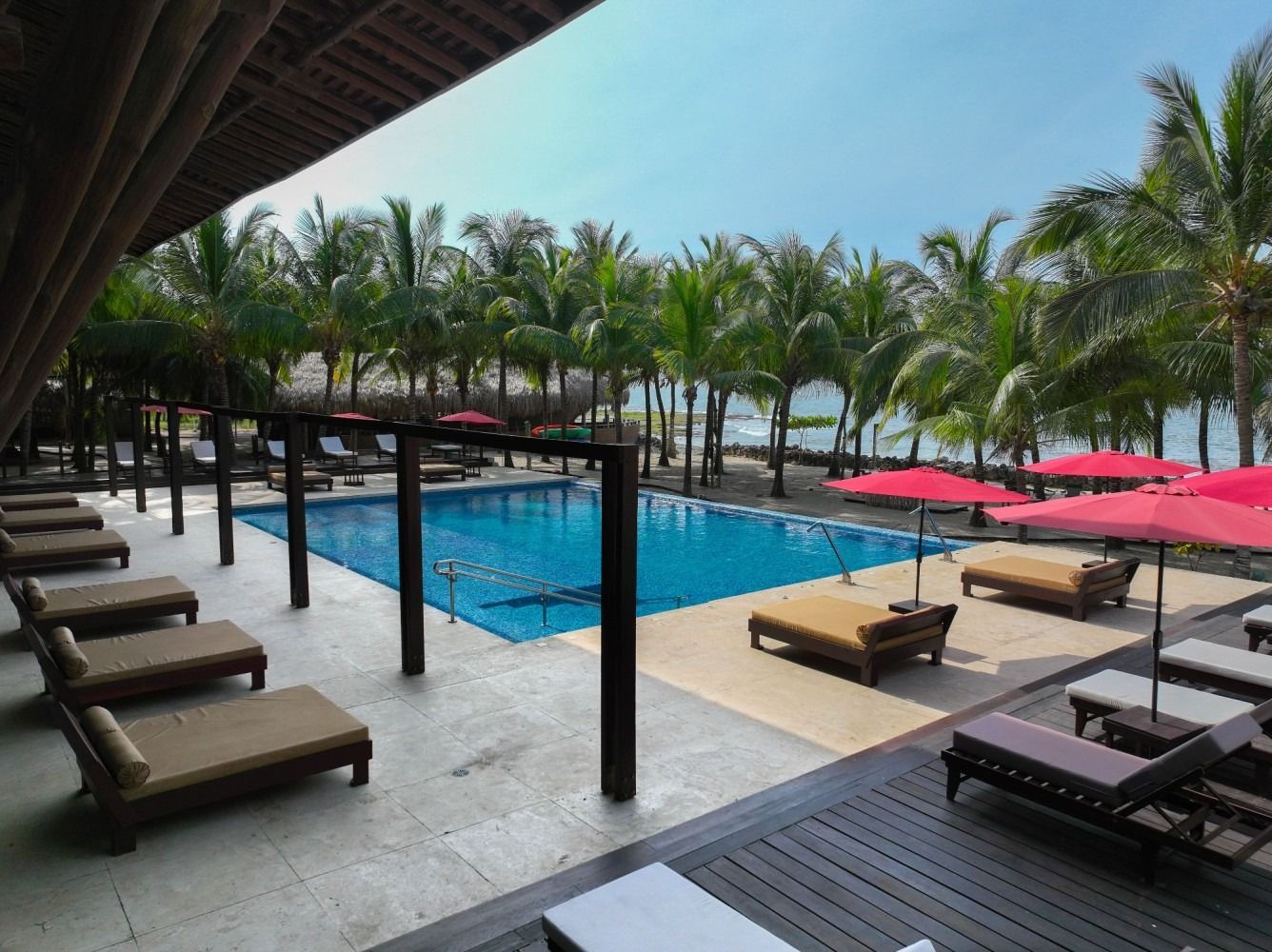 Swimming pool area with lounge chairs, red umbrellas, and palm trees, near the beach.