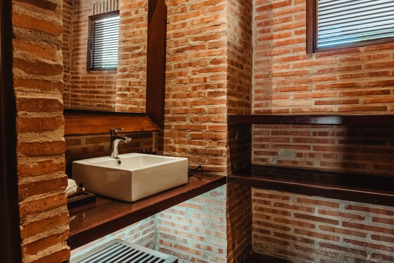 Brick-walled bathroom with a white rectangular sink on a dark wood shelf, dark wood accents, and a small window.