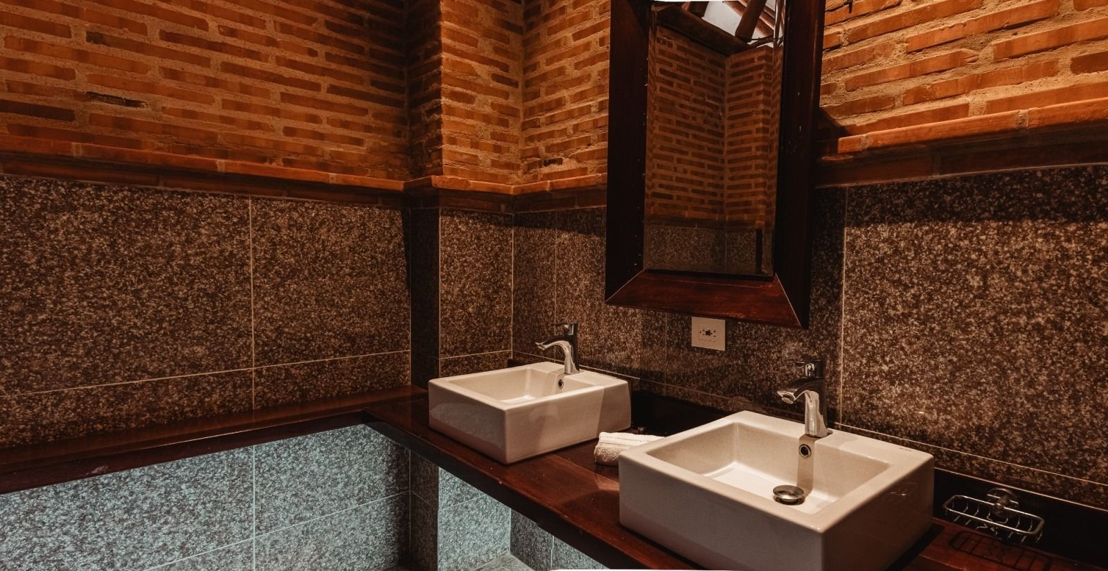 Bathroom with two white square sinks on a dark wood counter. Red brick and dark speckled walls. Mirror.