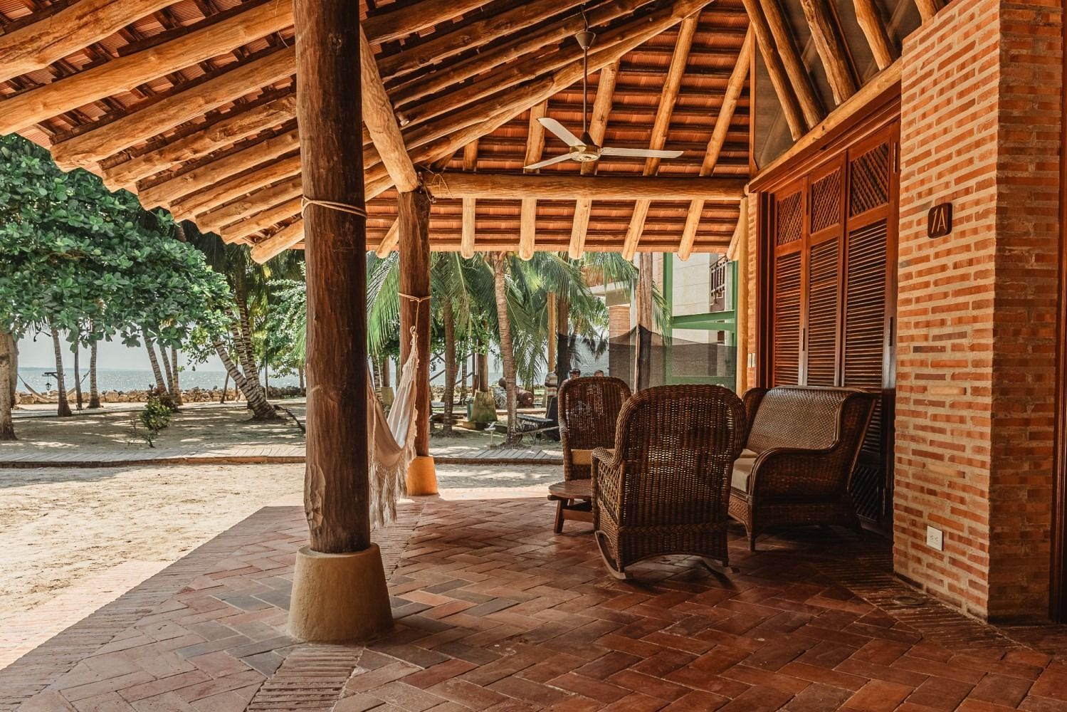 Covered patio with brick flooring, wooden beams, chairs, and view of beach and trees.
