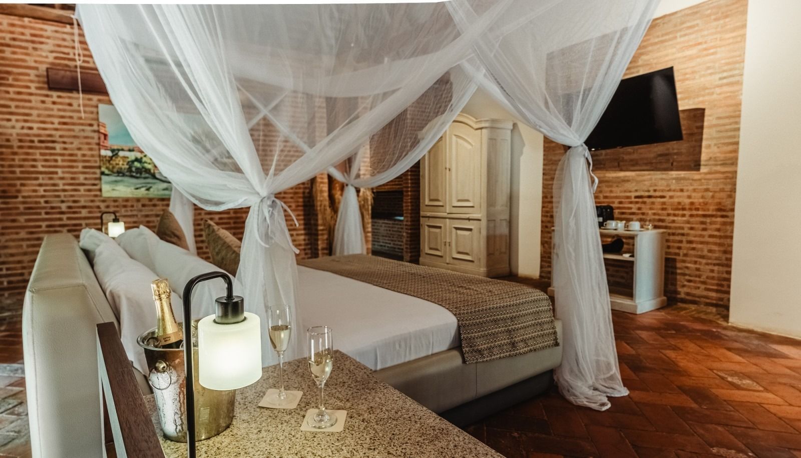Bedroom with canopy bed, exposed brick wall, champagne and glasses on a side table.