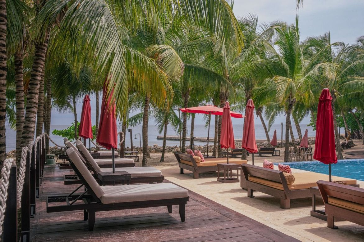 Lounge chairs and umbrellas by a pool and beach, palm trees overhead, red umbrellas, sunny setting.