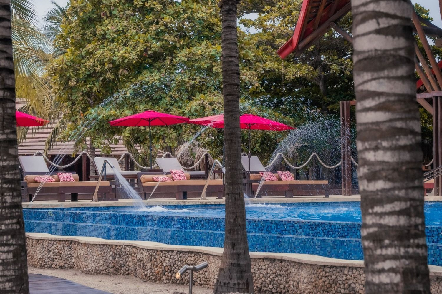Poolside scene: blue pool, water fountains, red umbrellas, lounge chairs, palm trees.