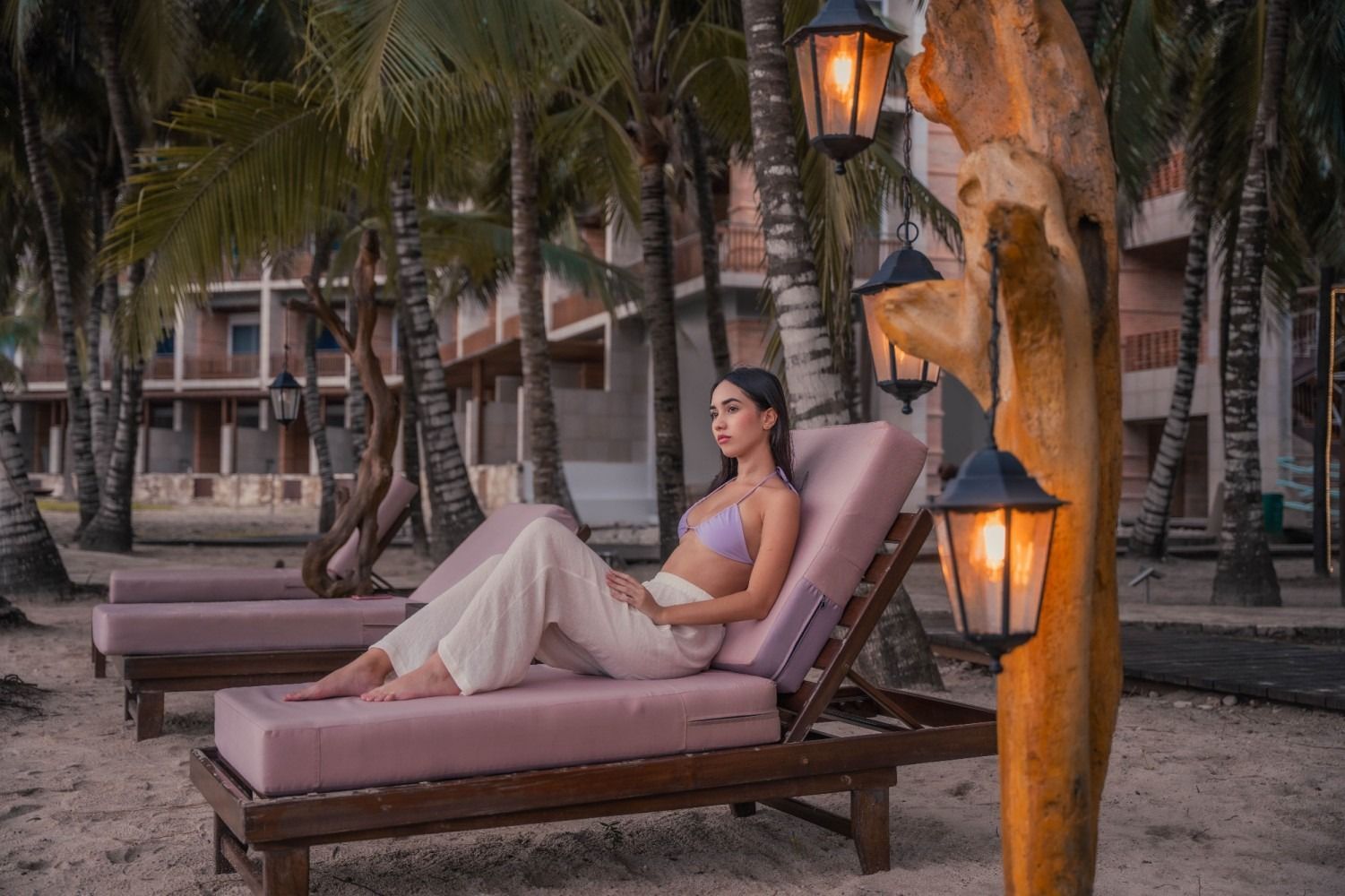 Woman relaxing on a pink sun lounger by the beach, under lanterns.