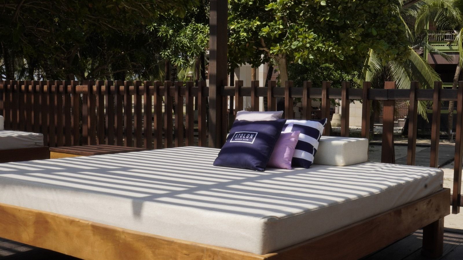 Wooden daybed with pillows, outdoors near a wooden fence and trees, bright sunlight.