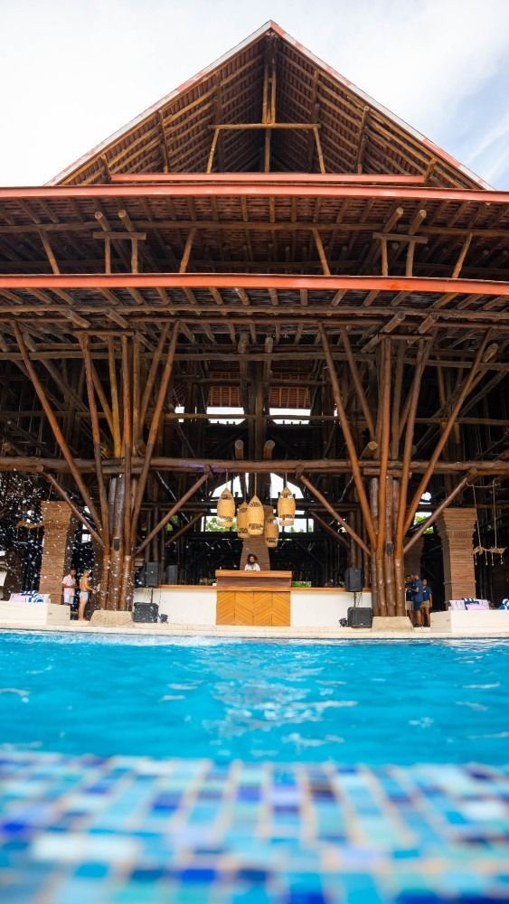 Large bamboo structure over a pool. Brown wood, blue water, and a cloudy sky.