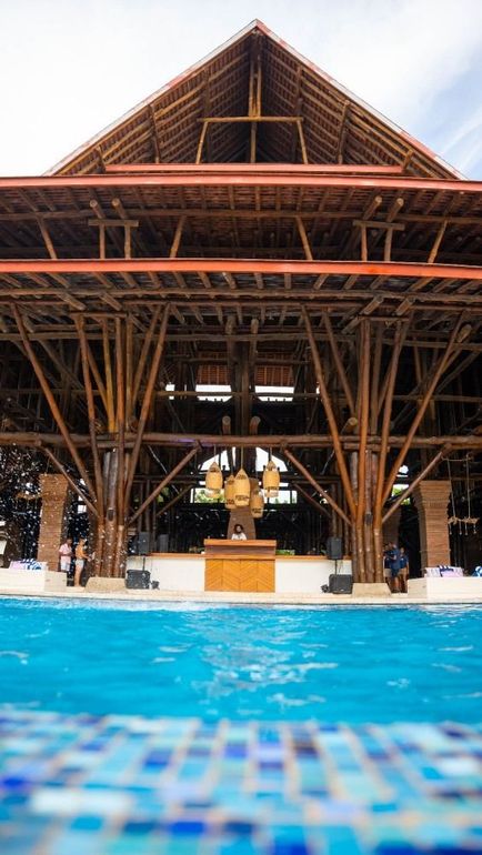 Large bamboo structure over a pool. Brown wood, blue water, and a cloudy sky.