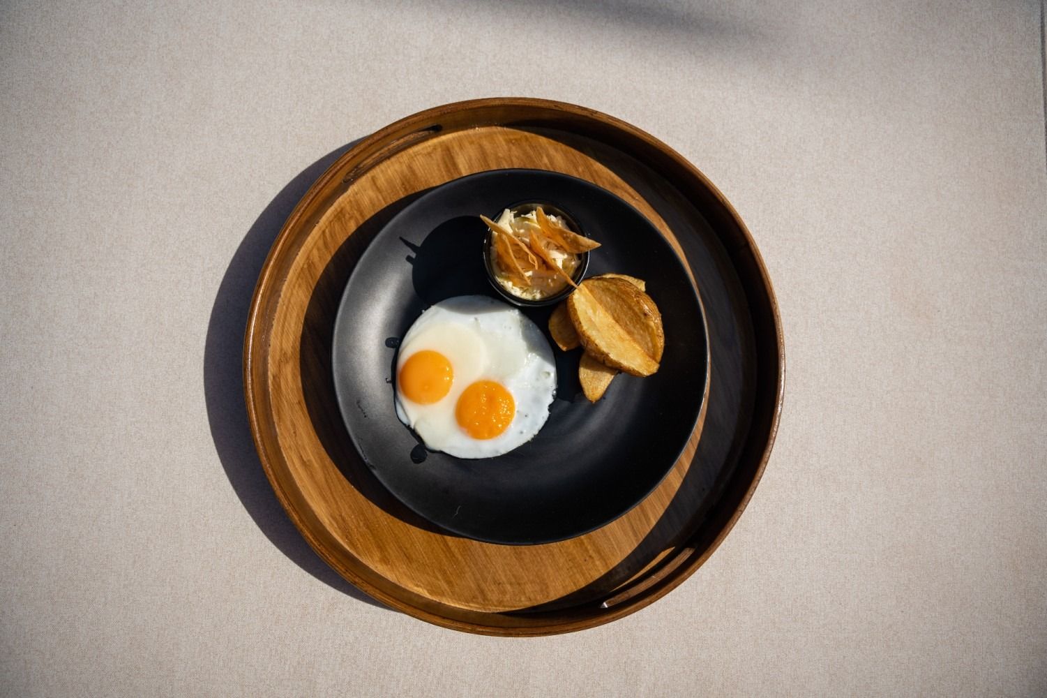 Two sunny-side-up eggs, potatoes, and an unknown food item on a black plate, presented on a wooden tray.