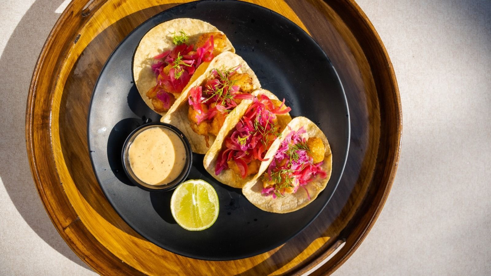 Tacos with pink slaw on a black plate, with sauce and lime on a wooden tray.