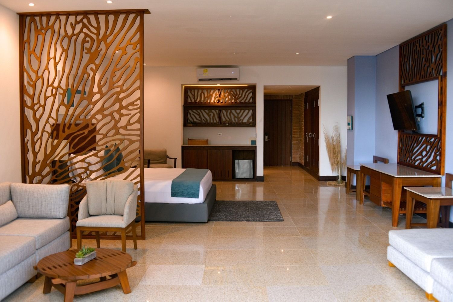 Hotel suite interior: wood screens, light-colored furniture, bed, TV, and bar area, with a beige tiled floor.