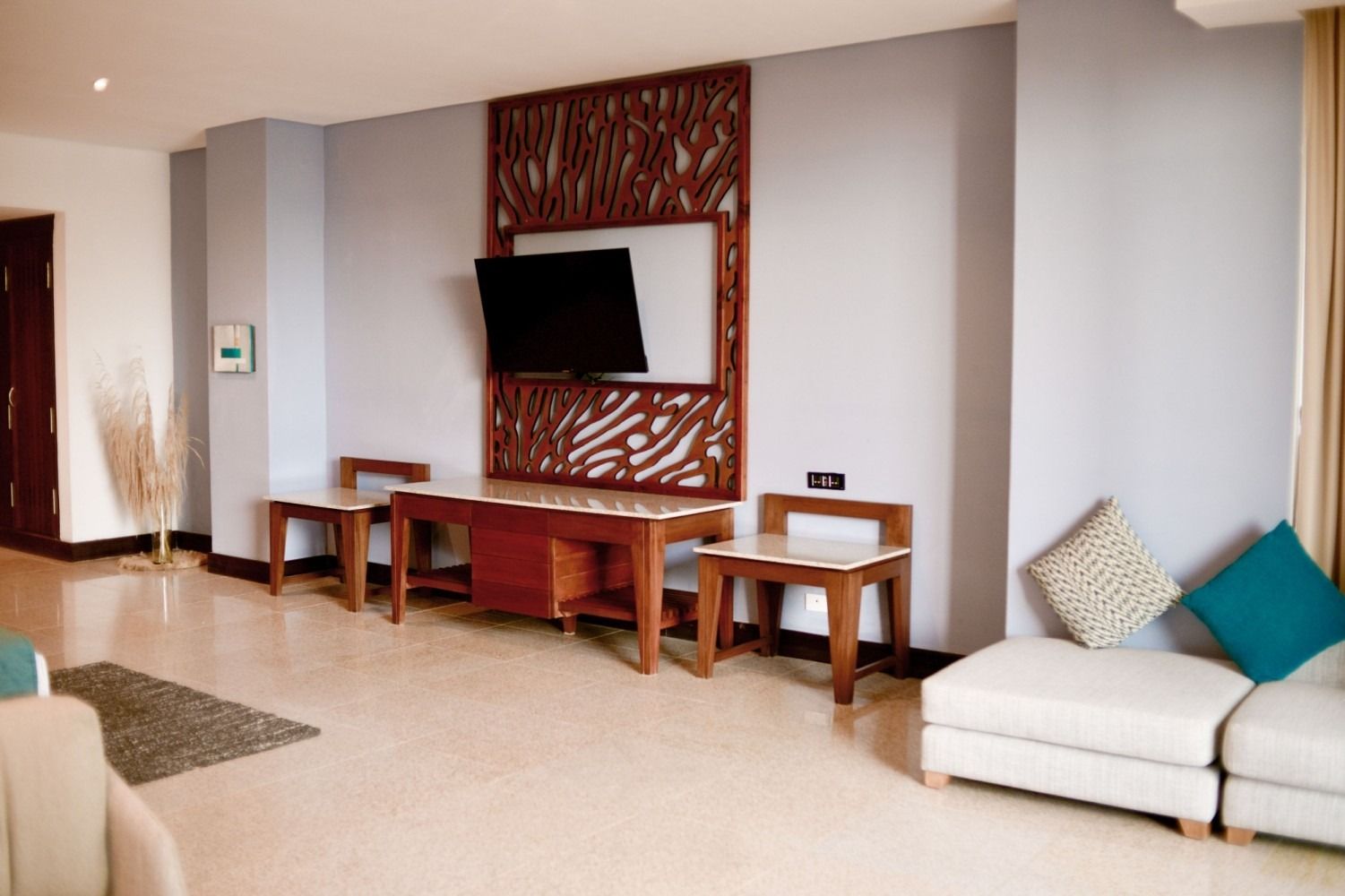 Living room with TV mounted on a decorative wooden panel, sofa, tables, and beige flooring.